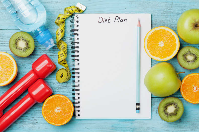饮食计划菜单旁边的卷尺哑铃和蓝色背景上的新鲜水果减肥食品