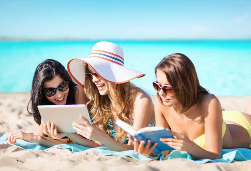 趴在沙滩上看着书本平板电脑的三个度假的年轻人