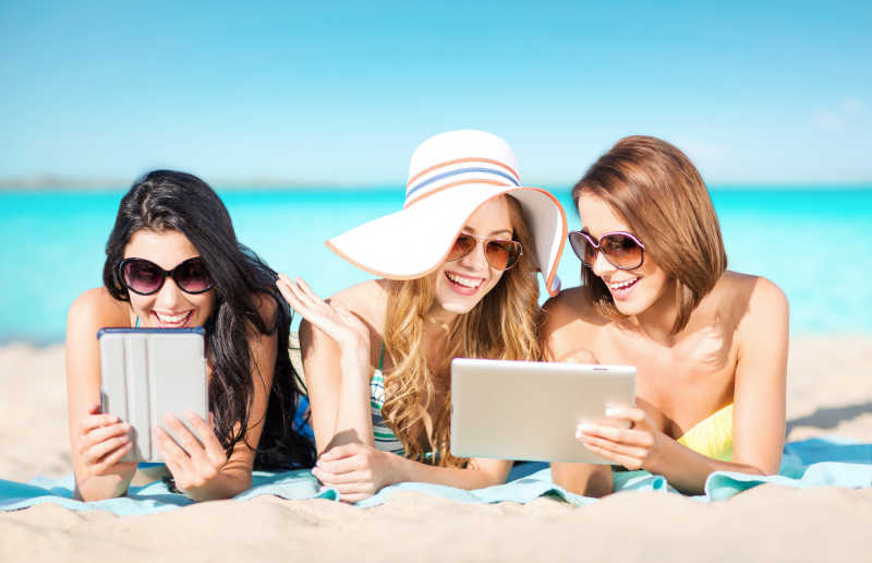 在海边沙滩上趴着的三个年轻美女看着平板电脑