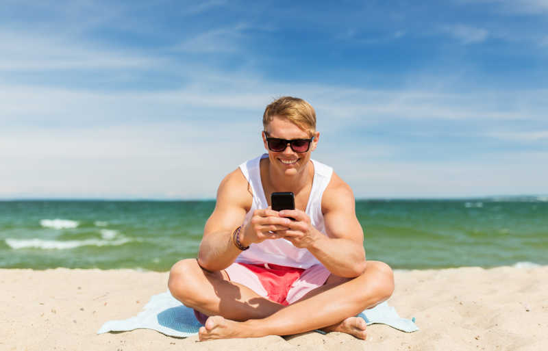 海滩上拿着智能手机的愉快微笑的年轻人