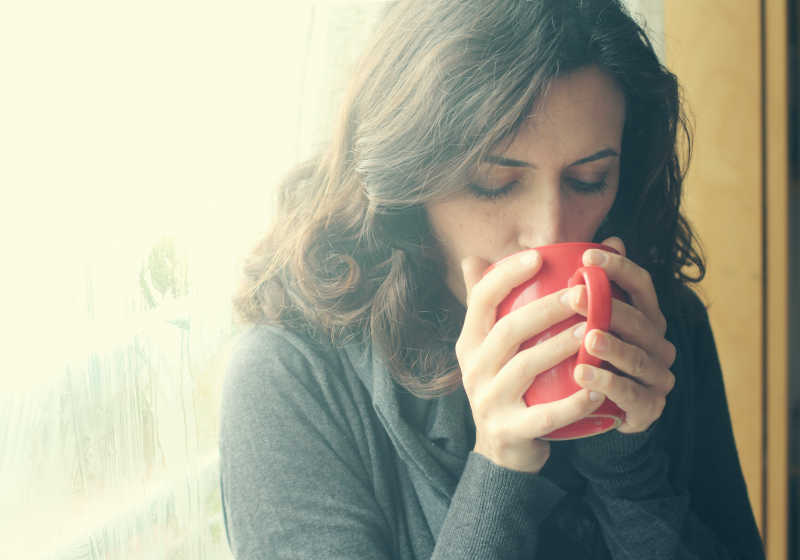年轻漂亮的女人站在窗边喝着一杯咖啡