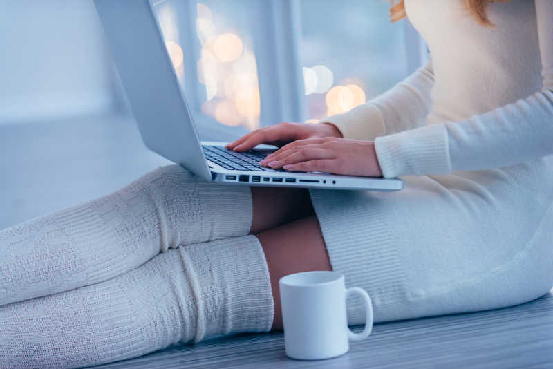 穿着白色毛衣和袜子坐在家里地上在笔记本电脑上工作旁边还有咖啡杯