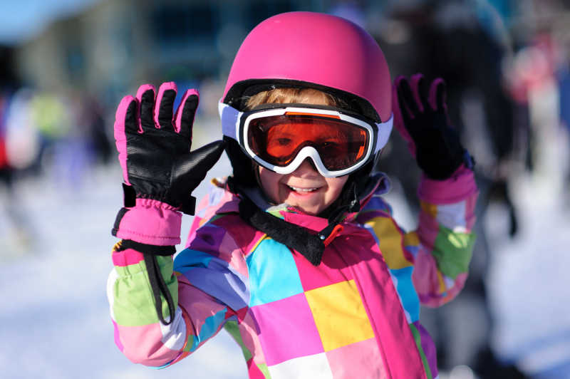 小女孩穿着色彩鲜艳的外套和一个粉红色的滑雪头盔和护目镜站在雪地里