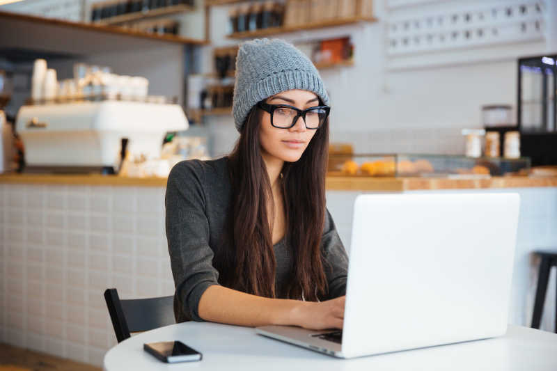 带着帽子和黑框眼镜的美女在咖啡厅使用笔记本电脑工作