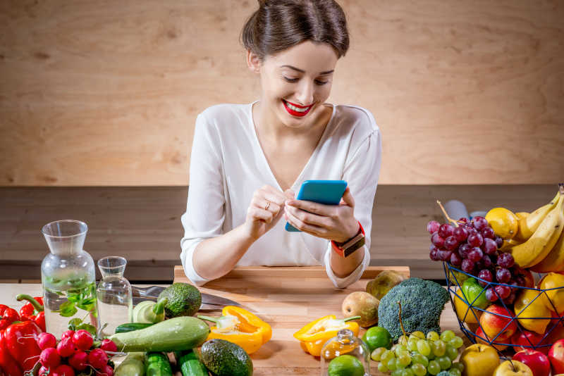 微笑看手机的美女在盛满水果蔬菜的餐桌上