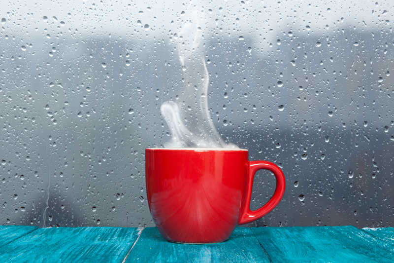 雨天背景下蓝色木质桌上的红色杯子里的热饮冒着蒸汽