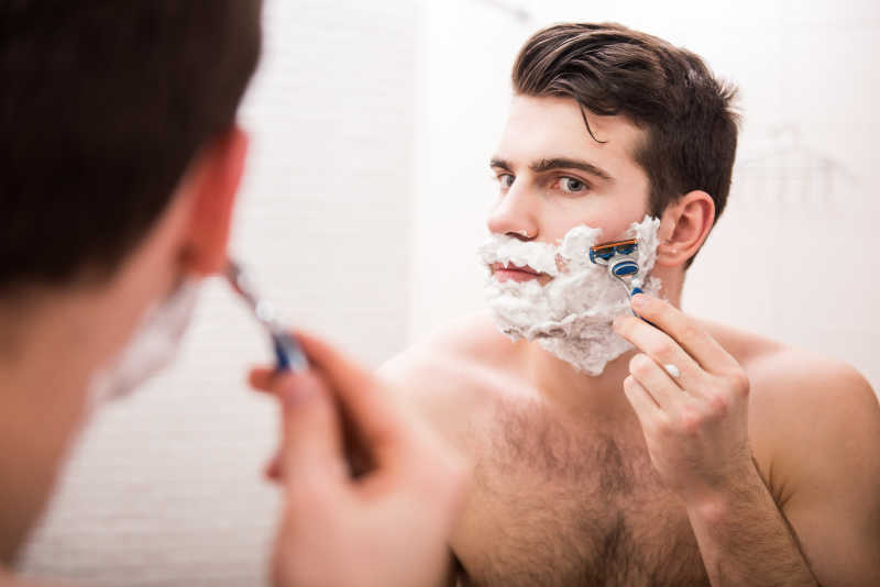 脸上抹着剃须泡沫的男人在用剃须刀刮胡子