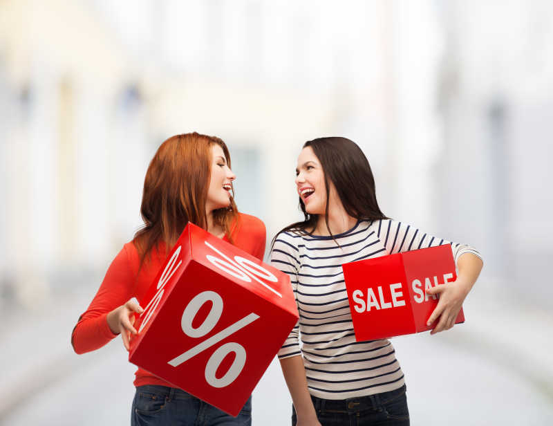 虚化街边背景下两个微笑的女人抱着写着销售字母的红盒子