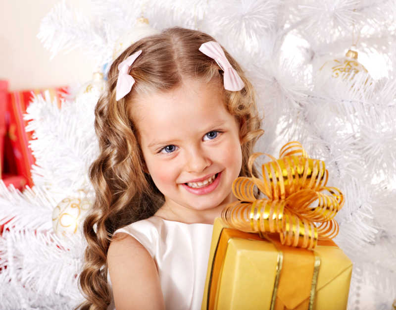 白色圣诞树旁抱着礼物微笑的小女孩