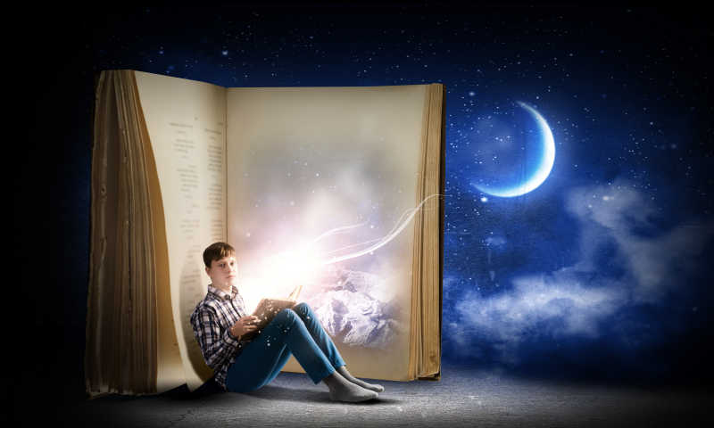 蓝色夜空下倚靠在巨大书籍上看书的小男孩
