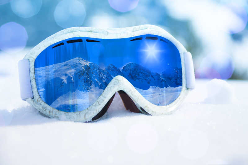 光斑背景下雪地上的滑雪护目镜倒映出的雪山