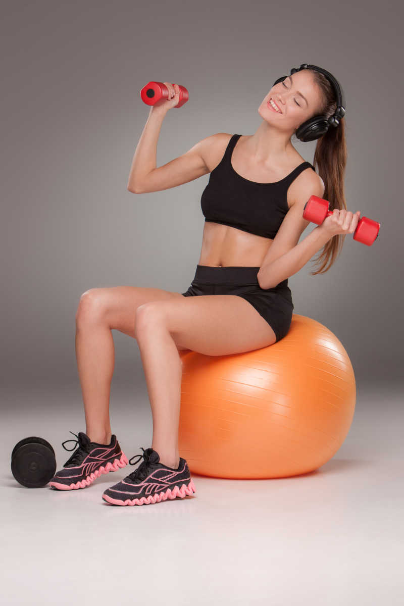 运动的女人做有氧运动与红色哑铃和耳机在健身球上灰色的背景