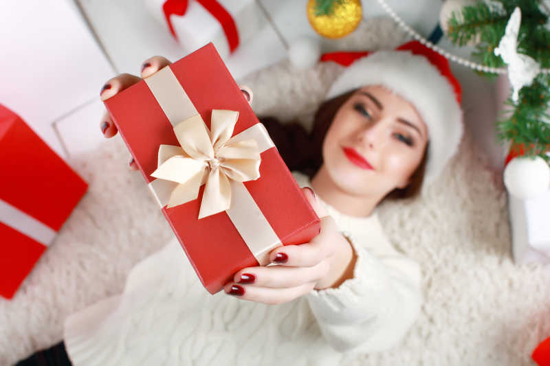戴着圣诞帽子的年轻女子躺在白色地毯上手拿礼品盒