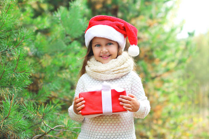 带着圣诞帽子和白色围巾的小女孩手拿红色礼品盒