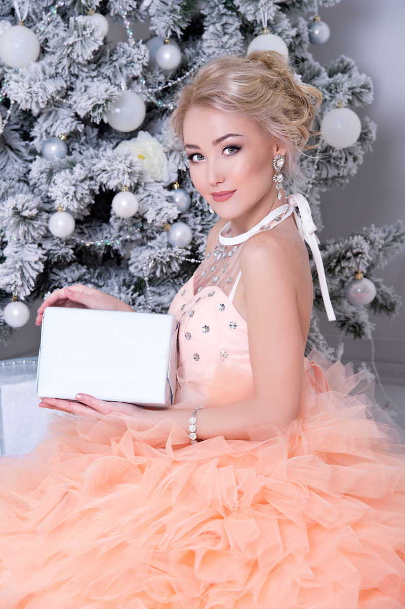美丽的圣诞树前穿着粉色长裙的金发美女手拿白色礼品盒