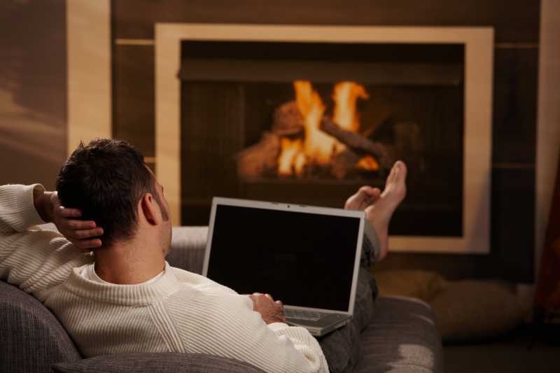 男人坐在家里的壁炉前的沙发上使用笔记本电脑