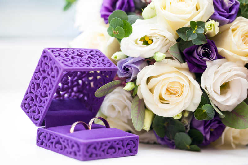 在一束鲜艳的玫瑰花旁边的紫色盒子里放着一对戒指