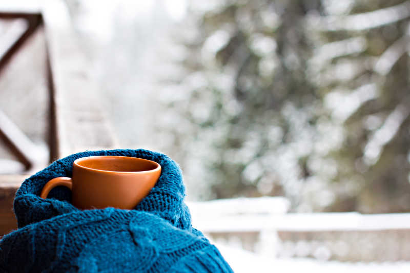冬季户外景观背景下蓝色针织衫包裹着一只橘色的杯子