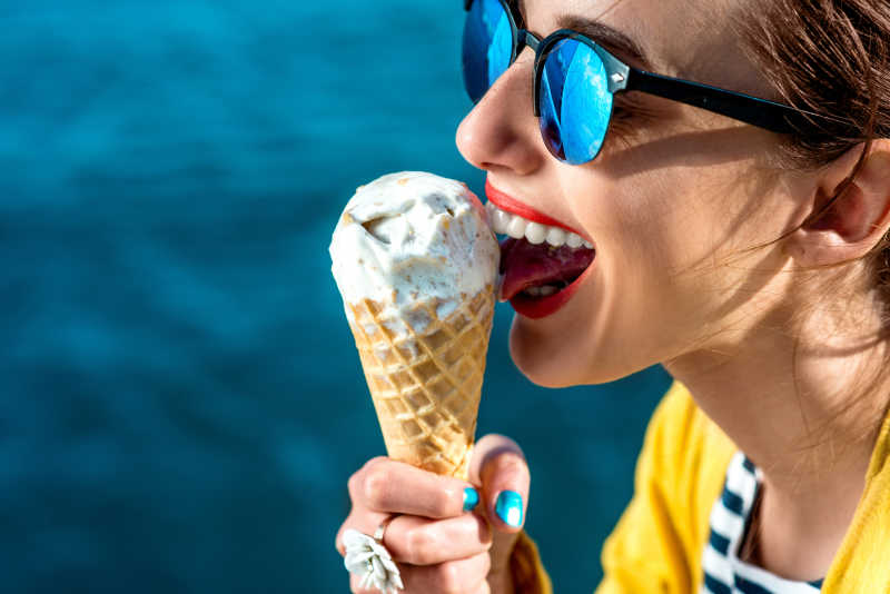 穿着黄色毛衣戴着太阳镜的年轻女子在蓝色的水面上吃冰淇淋