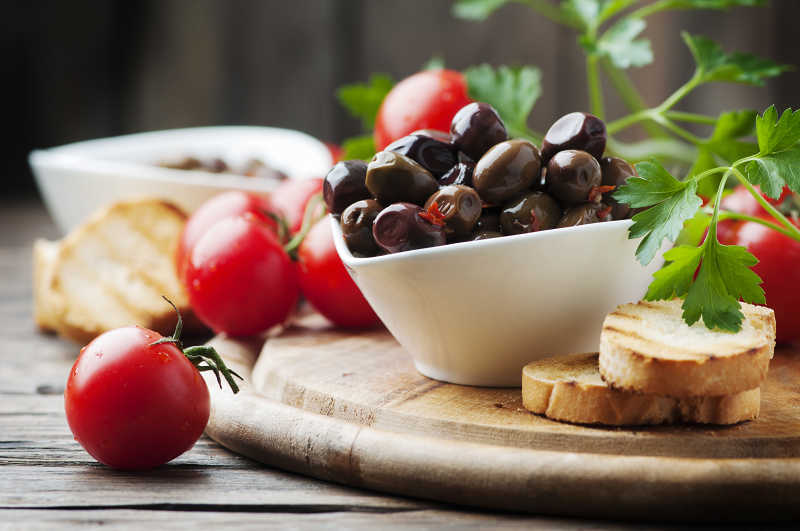 木桌上瓷碗中满满的橄榄和散落一旁的西红柿和馒头干