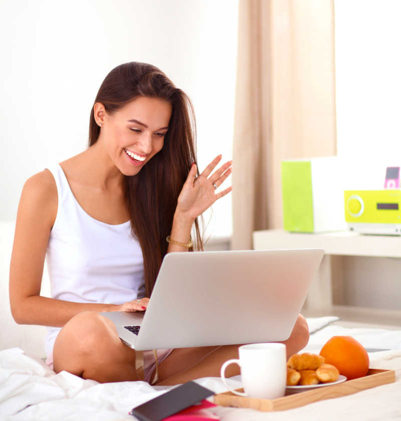 年轻漂亮的女人坐在床上使用笔记本电脑身边放着食物和手机