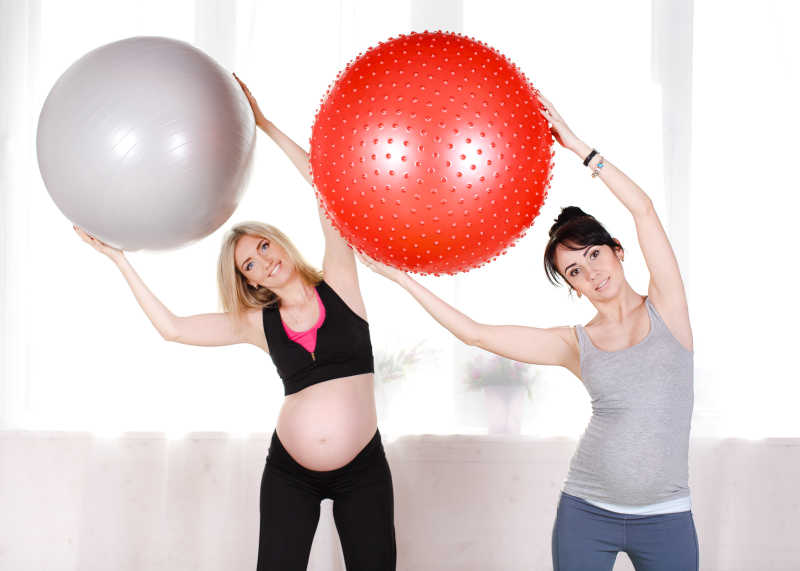 双手高举瑜伽球练习健身的金发和黑发孕妇