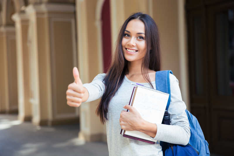 漂亮的女学生背着书包抱着书竖着大拇指