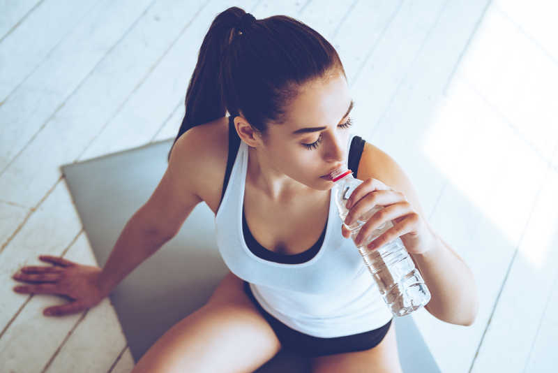 美丽的年轻女子坐在健身房运动垫上喝水