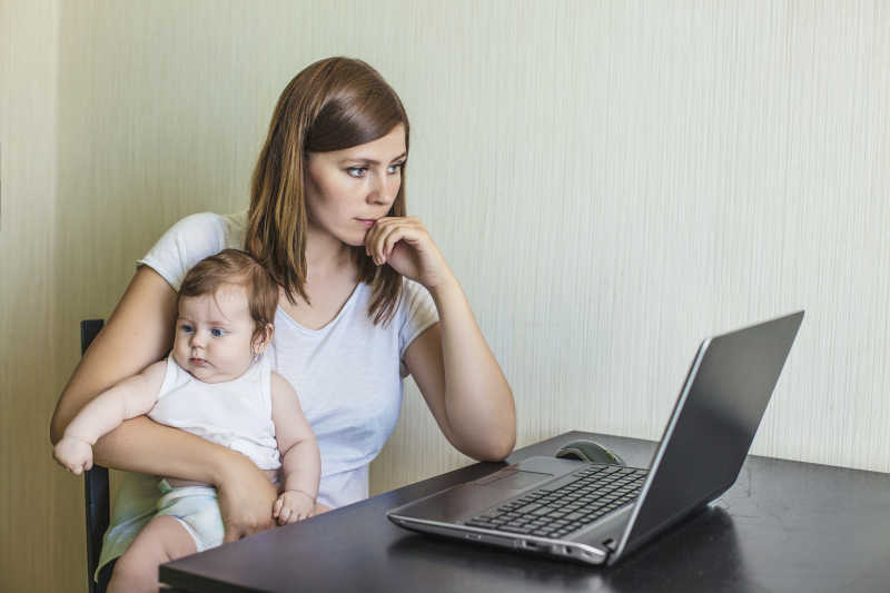 美女妈妈抱着宝宝坐在办公桌上的笔记本电脑前工作