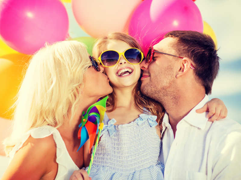 五颜六色的气球背景前幸福的父母亲吻小女孩