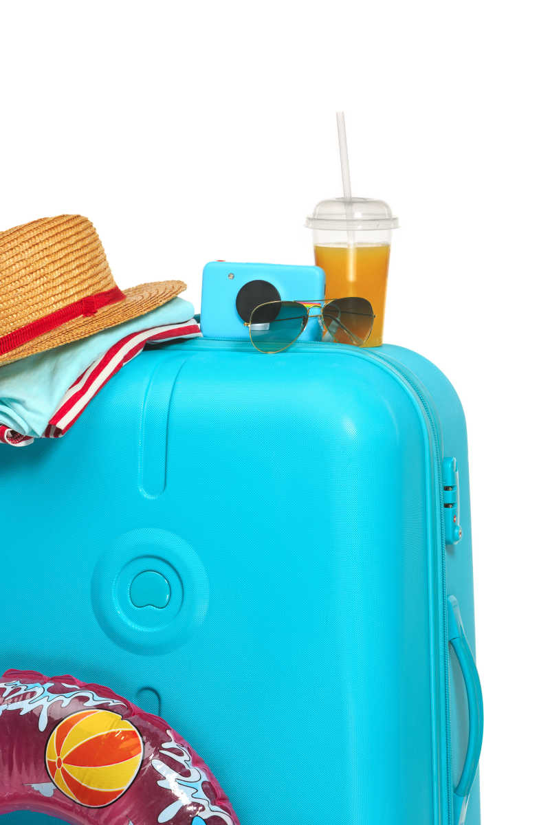 白色背景下放着遮阳帽太阳眼镜饮料和照相机的蓝色行李箱