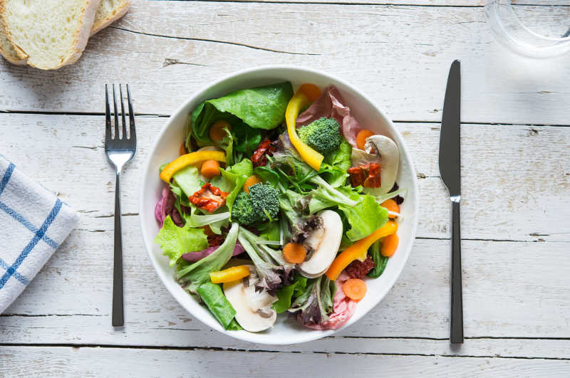 木桌上的刀叉和白色碗中美味可口的蔬菜沙拉