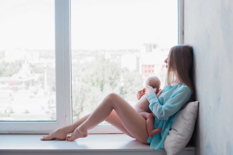 抱着小婴儿坐在白色飘窗台的年轻母亲
