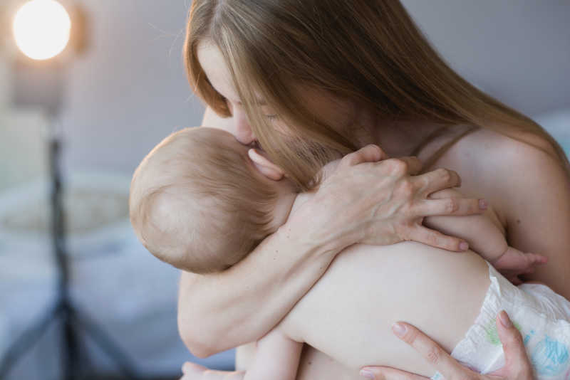 灰色有灯光的背景下抱着小婴儿亲吻的年轻妈妈