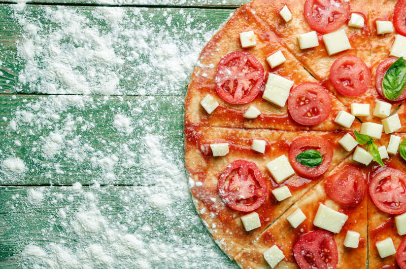 撒着面粉的绿色木桌上放着新鲜美味的切好的西红柿披萨