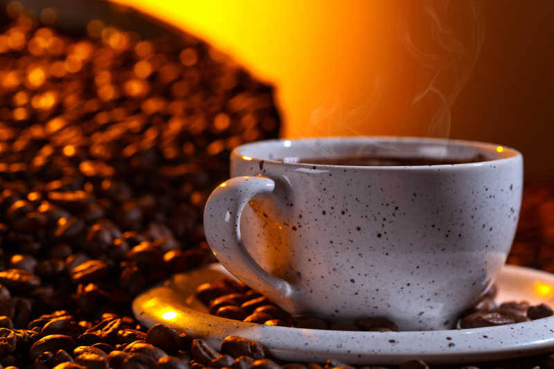 金色朝阳下的冒着热气的一杯咖啡和棕色的咖啡豆