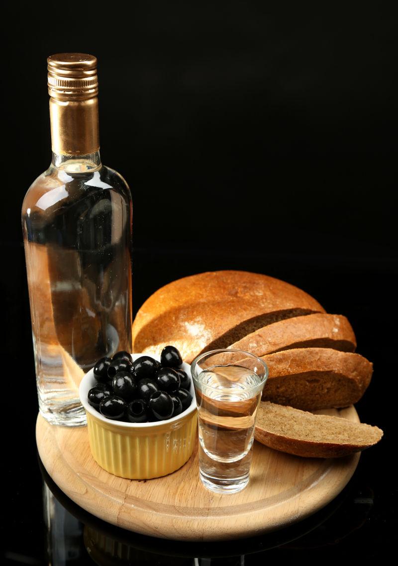 黑色背景下的面包与一瓶伏特加酒和腌制橄榄
