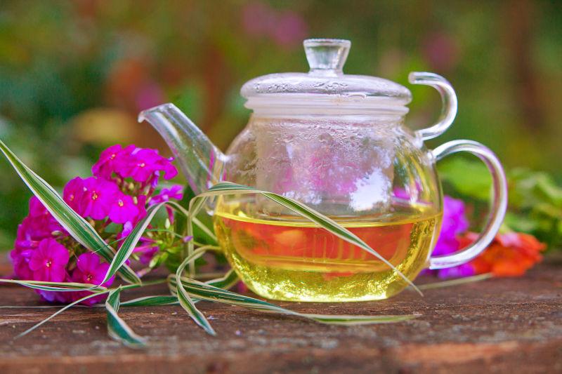 木桌上粉色鲜花旁边的盛着绿茶的玻璃茶壶