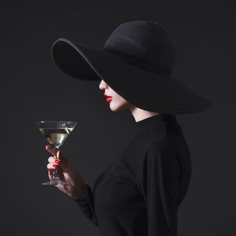 在黑色背景下戴着大黑帽子的豪华女人手里拿着一杯马蒂尼酒