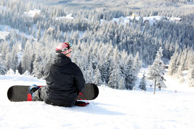落满雪的松树林背景下的抱着单板坐在雪地上休息的人