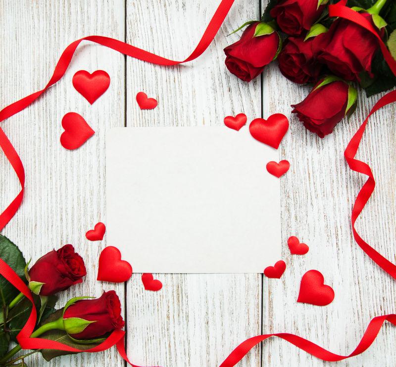 木板上空白纸张周围装饰着美丽的玫瑰花束和红色丝带还有爱心饰品