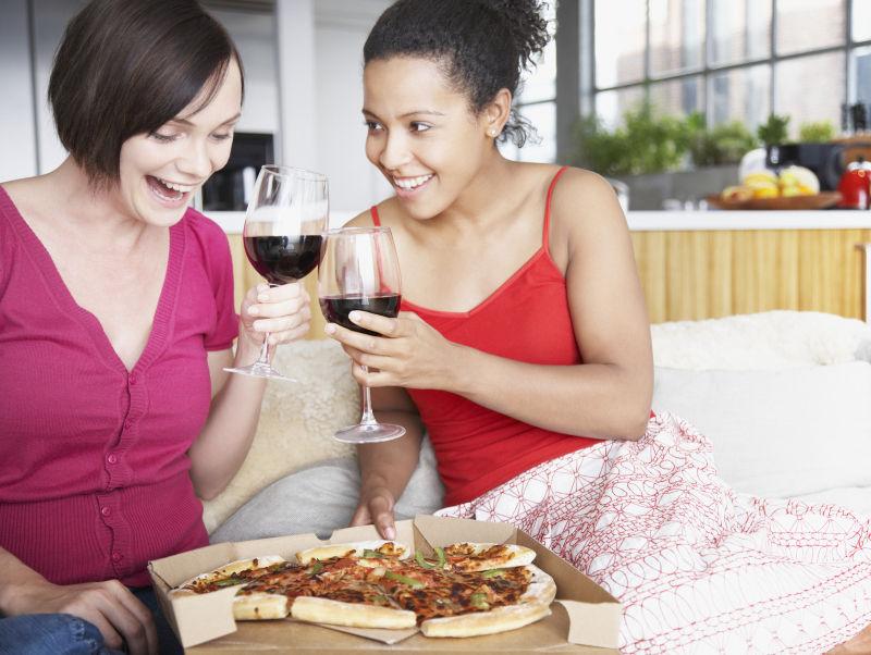 在室内的沙发上吃着披萨喝着红酒的两个女人
