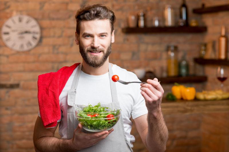 穿着白色T恤和围裙的漂亮胡子男人在厨房里吃西红柿沙拉
