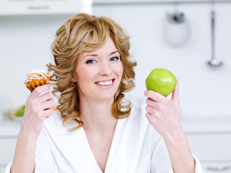 快乐的年轻漂亮的女人坐在厨房里拿着绿色的苹果和甜的蛋糕