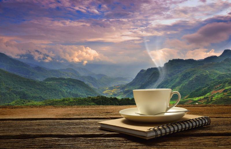 蓝天白云和群山背景下的日记本上的一杯热咖啡