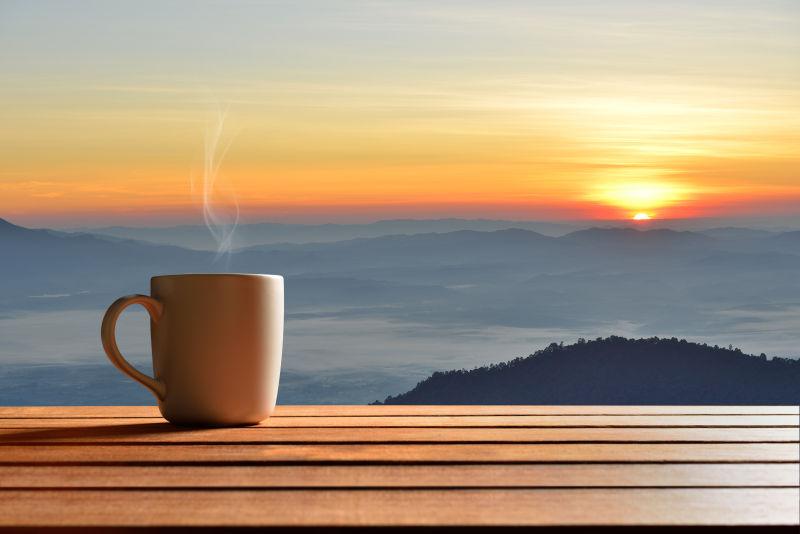 群山上的日出背景下的木桌上的一杯热咖啡