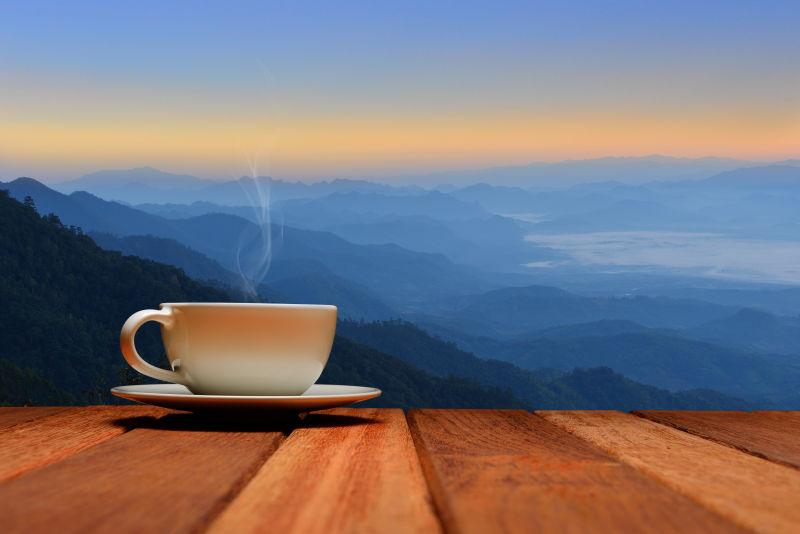 日出时分的群山背景下的木桌上的一杯热咖啡