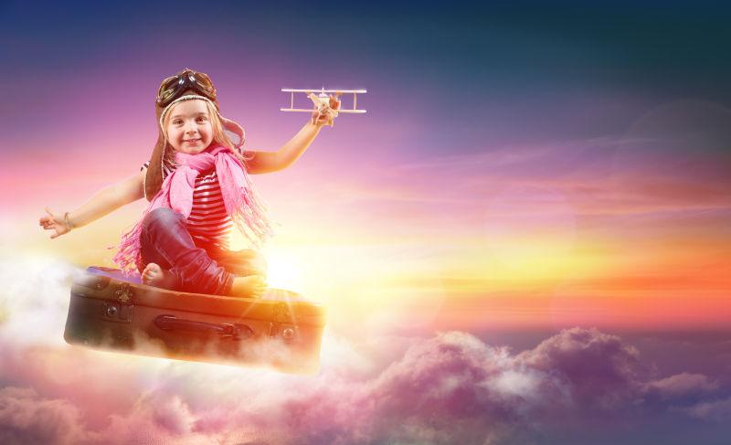 夕阳下飞在空中坐在行李箱上玩玩具飞机的小女孩