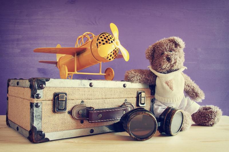 紫色背景下的行李箱上的玩具飞机和毛绒玩具熊