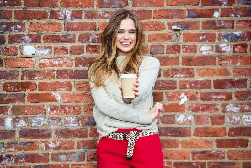 美丽的快乐女孩喝咖啡微笑着靠近红砖墙